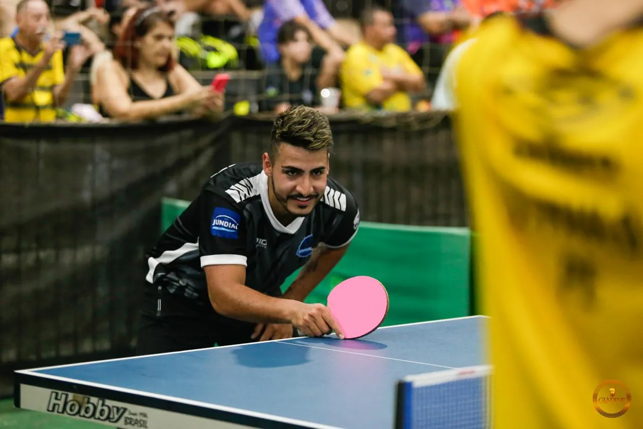 Jonathan sorrindo na mesa, pronto para sacar, durante um torneio com torcida ao fundo